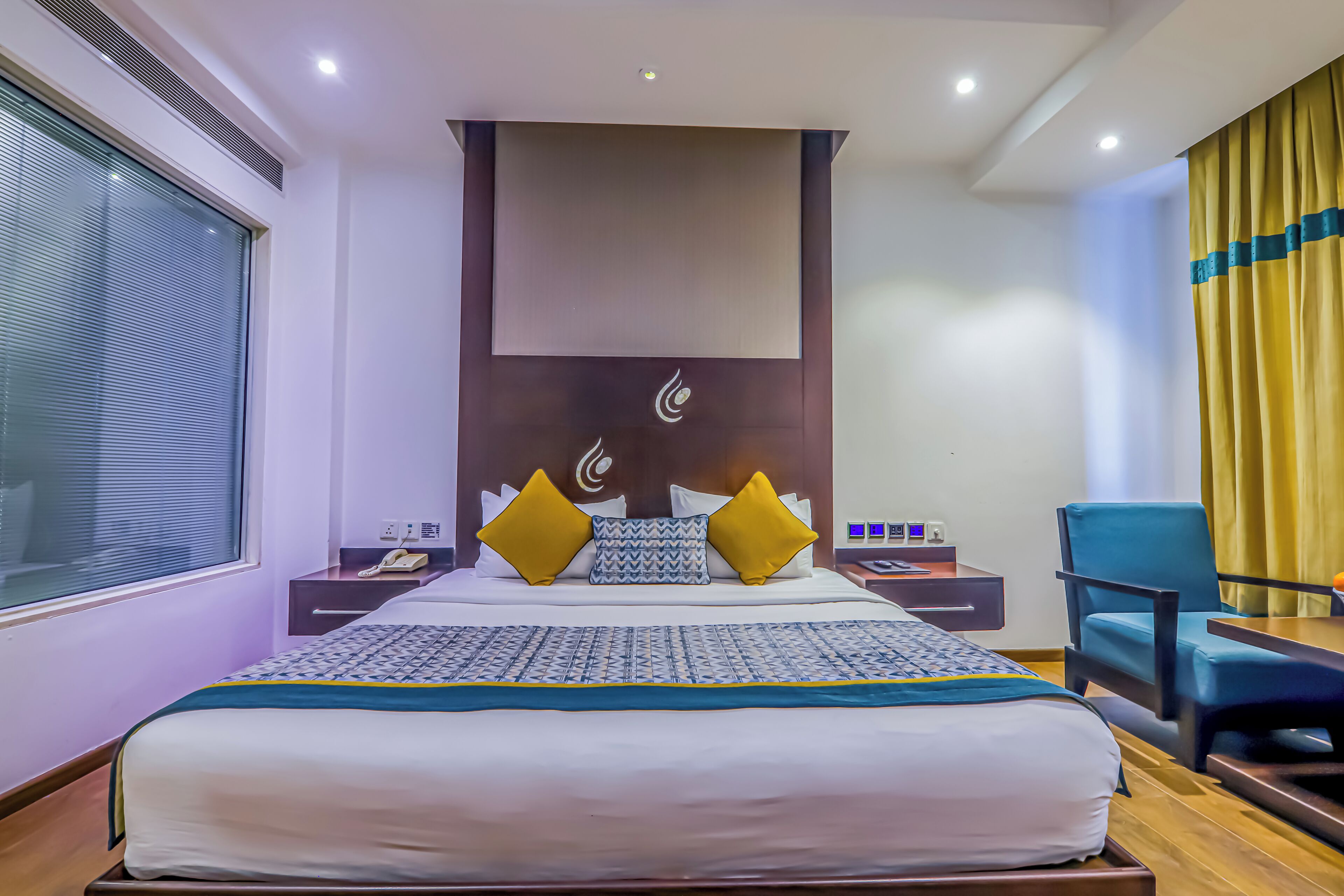 A hotel room with a large bed, a desk, a chair, and a window with blinds.
