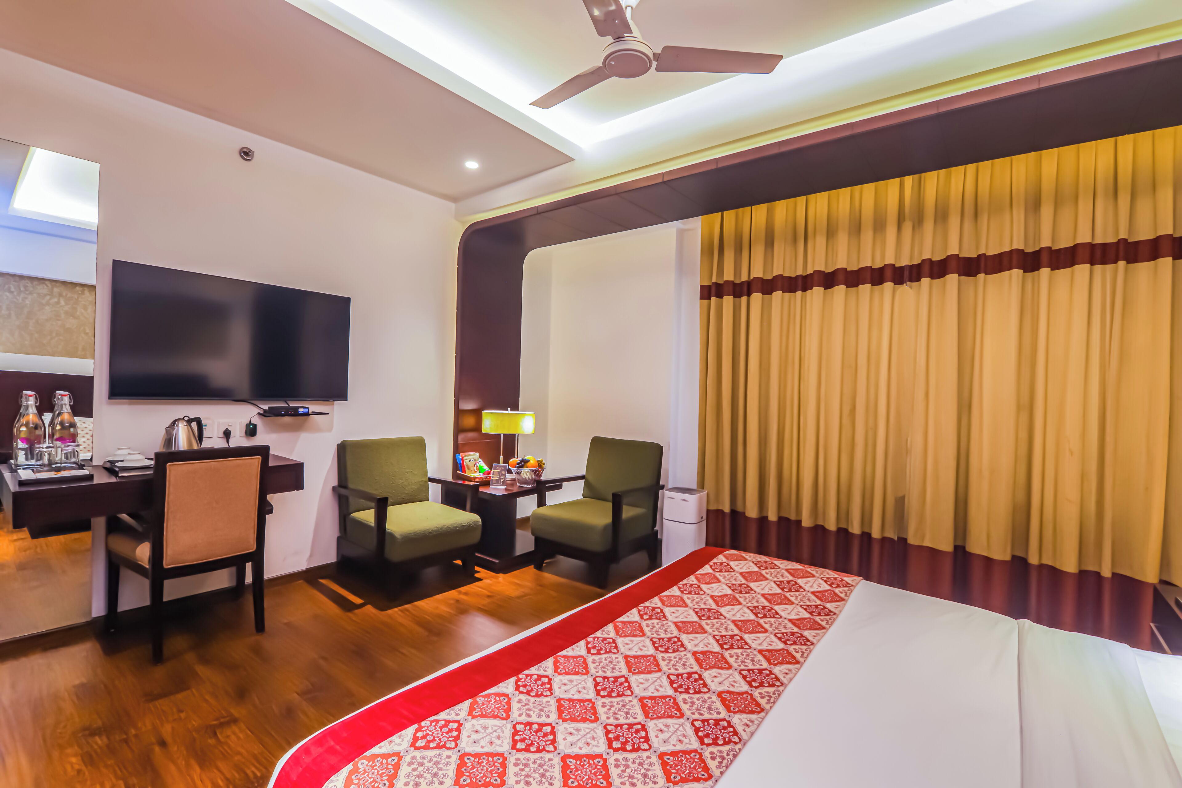 Habitación de hotel con una cama, dos sillas, un escritorio con televisor de pantalla plana, ventilador de techo y una ventana con cortinas.
