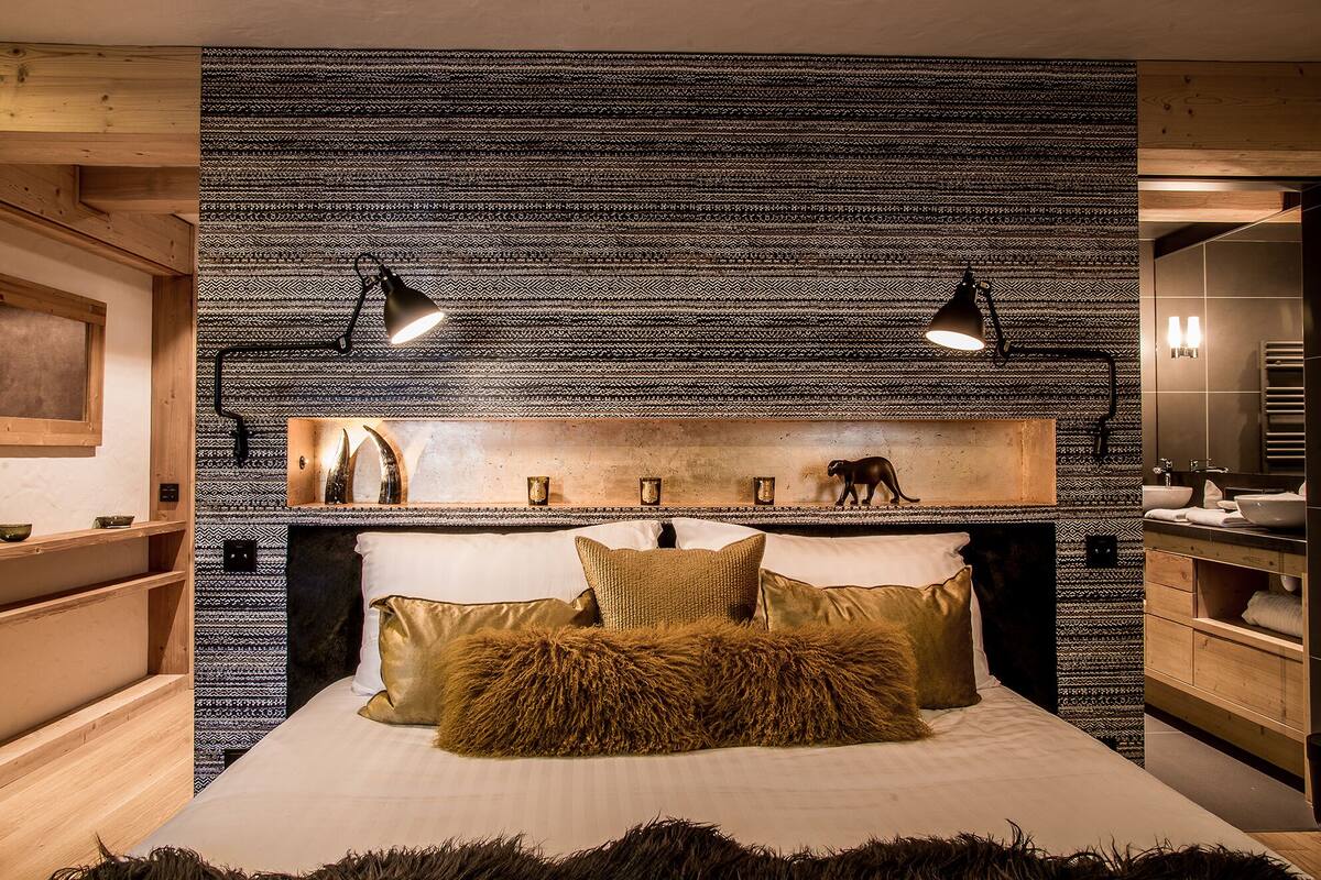 A bedroom with a patterned wall, a bed with pillows, a nightstand, and a mirror.