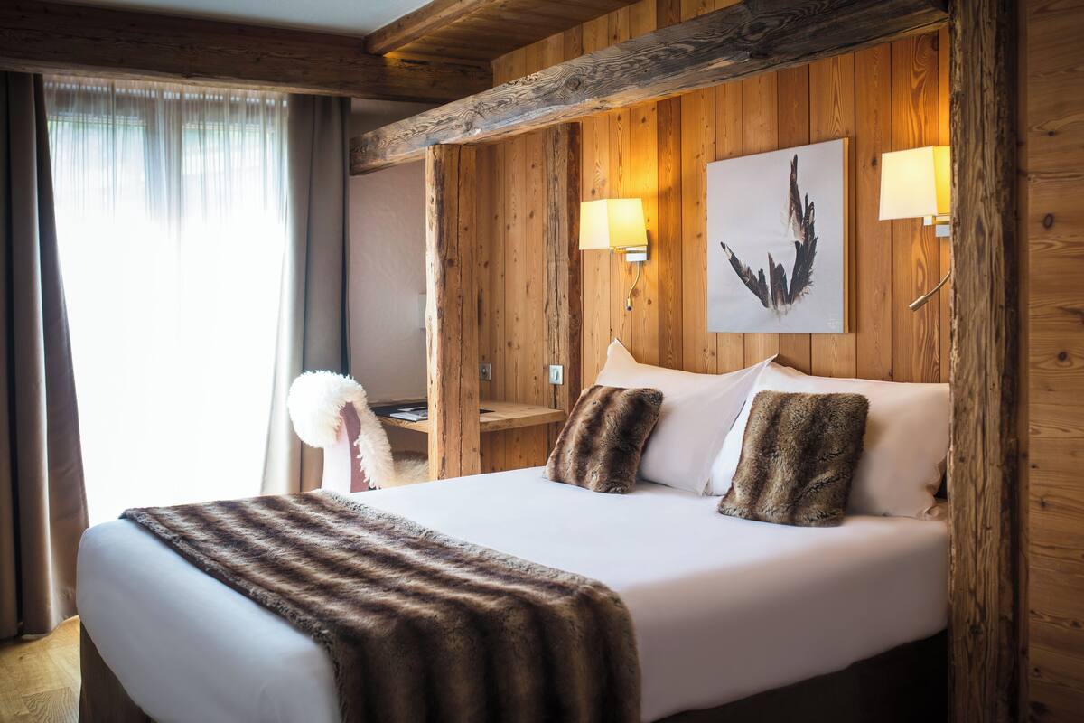 A hotel room with a wooden paneled wall, a bed with white linens, two wall-mounted lamps, and a framed artwork above the bed.