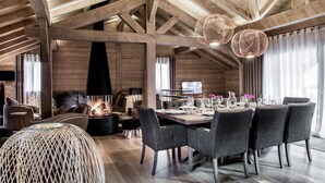 Una habitación amplia con techo alto, vigas de madera, chimenea, zona de comedor con mesa dispuesta para seis personas y un rincón de estar acogedor con sofá y sillones.