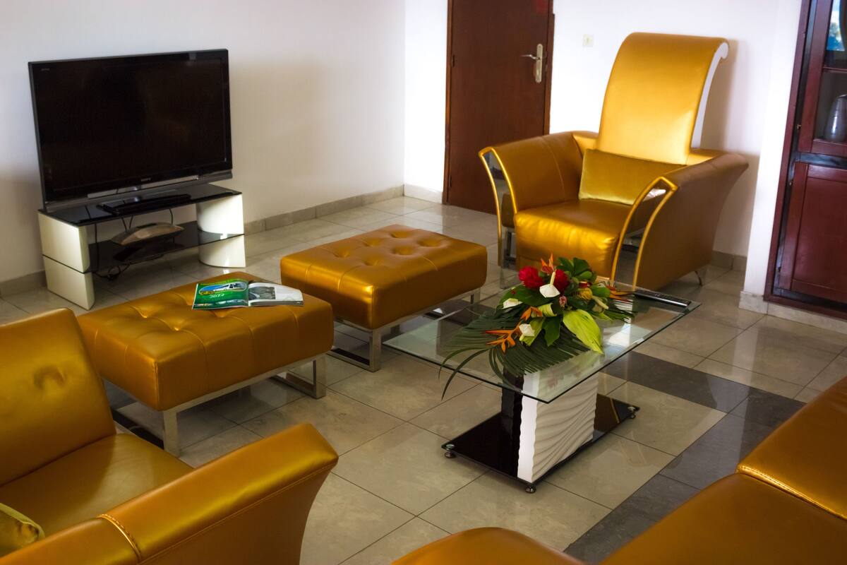 A modern living room with a flat-screen TV, a glass coffee table with a floral arrangement, and yellow leather furniture.