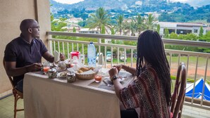 A balcony with a table set for two people, a water bottle, and a basket of bread. - Hotel Azur (Yaoundé)