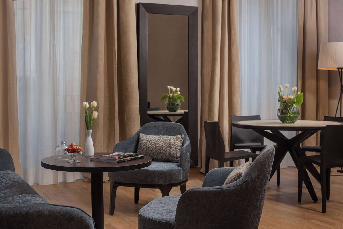 A modern hotel room with a round dining table, two grey armchairs, a small round table with a vase of flowers, and a large mirror on the wall.
