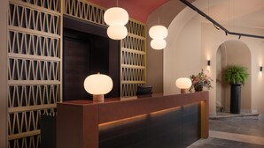 A reception desk with a modern design, featuring a dark wood finish and a geometric patterned wall behind it. There are three spherical pendant lights above the desk, and a potted plant is placed to the right. - NH Collection Palermo Palazzo Sitano (Palermo)