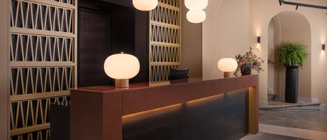 Un comptoir d’accueil au design moderne, avec un revêtement en bois foncé et un mur orné d’un motif géométrique en arrière-plan. Trois lampes suspendues sphériques sont disposées au-dessus du comptoir, et un pot de plante est placé sur la droite.
