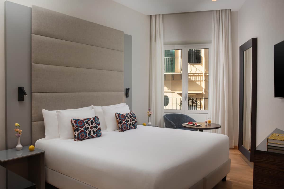 A hotel room with a large bed, a small table with a chair, a window with a view of a balcony, and a TV mounted on the wall.