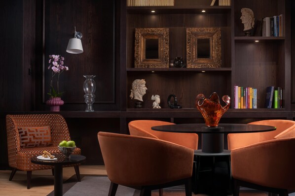 Una sala de estar moderna con un panel de pared de madera oscura, una mesa redonda, sillas anaranjadas, una mesita auxiliar con un cuenco de fruta, un jarrón con flores y una estantería con libros y objetos decorativos.