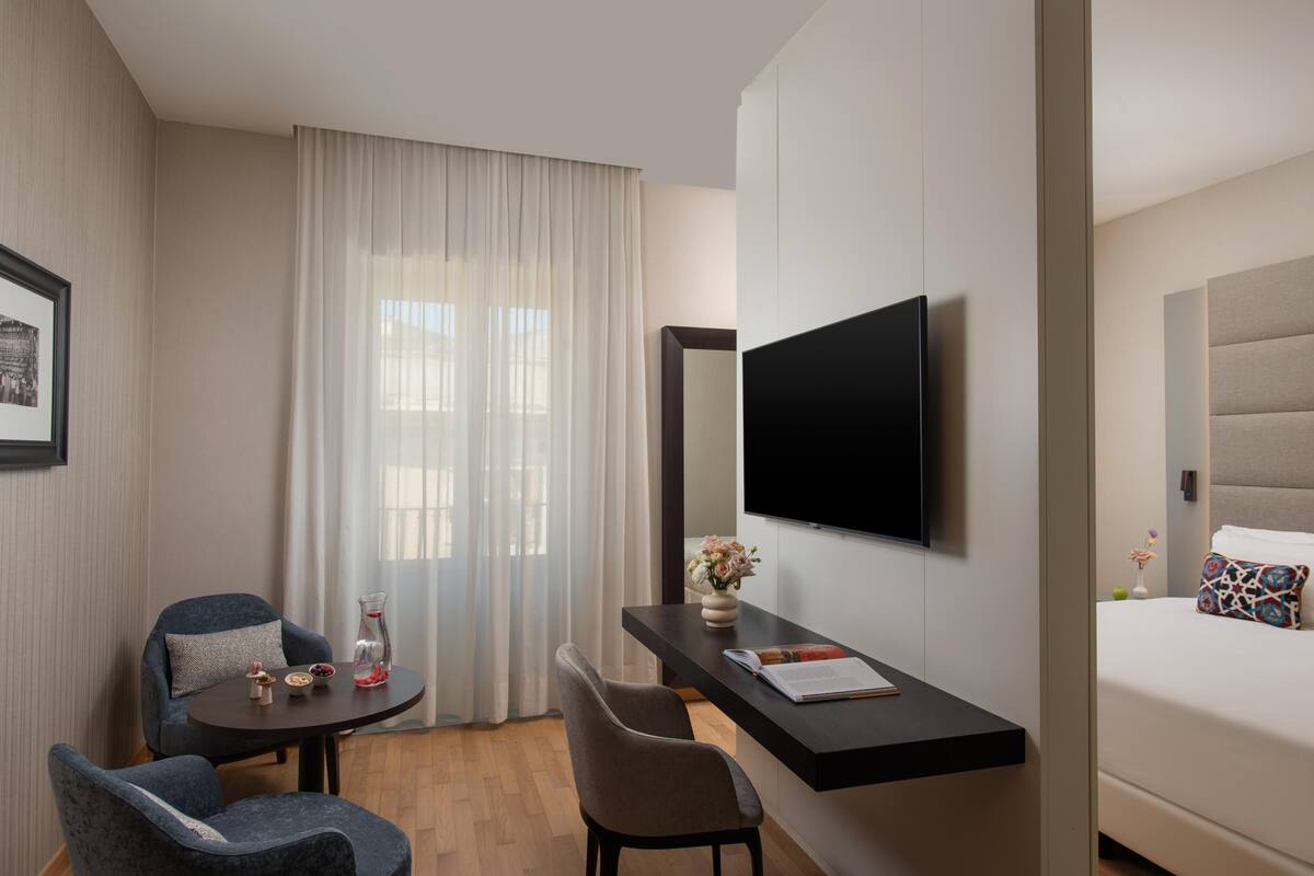A modern hotel room with a flat-screen TV mounted on the wall, a small round table with a vase of flowers, two chairs, and a bed with patterned pillows.
