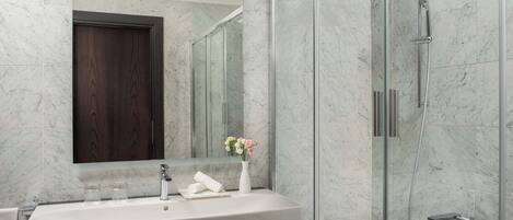 Un baño con pared de azulejos de mármol, puerta de madera, lavamanos blanco, espejo, cabina de ducha con mampara de cristal y bañera.