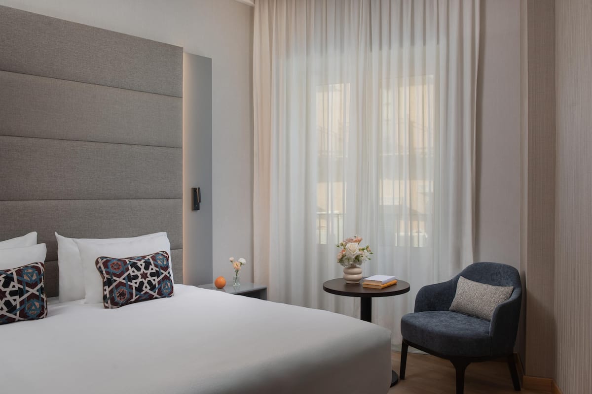 A hotel room with a large bed, a chair, a small table with a vase and book, and a window with sheer curtains.