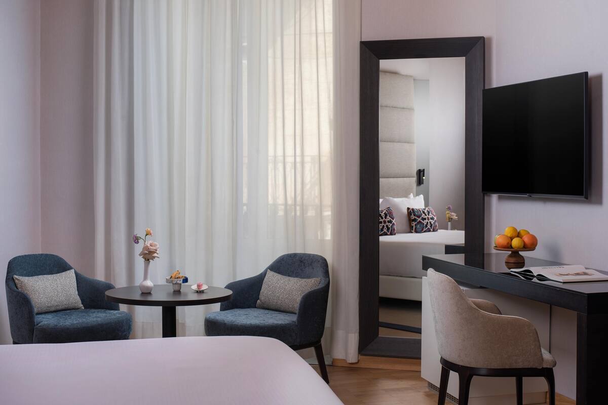 A hotel room with a bed, two armchairs, a small table with a vase, a desk with a bowl of fruit, and a TV mounted on the wall.