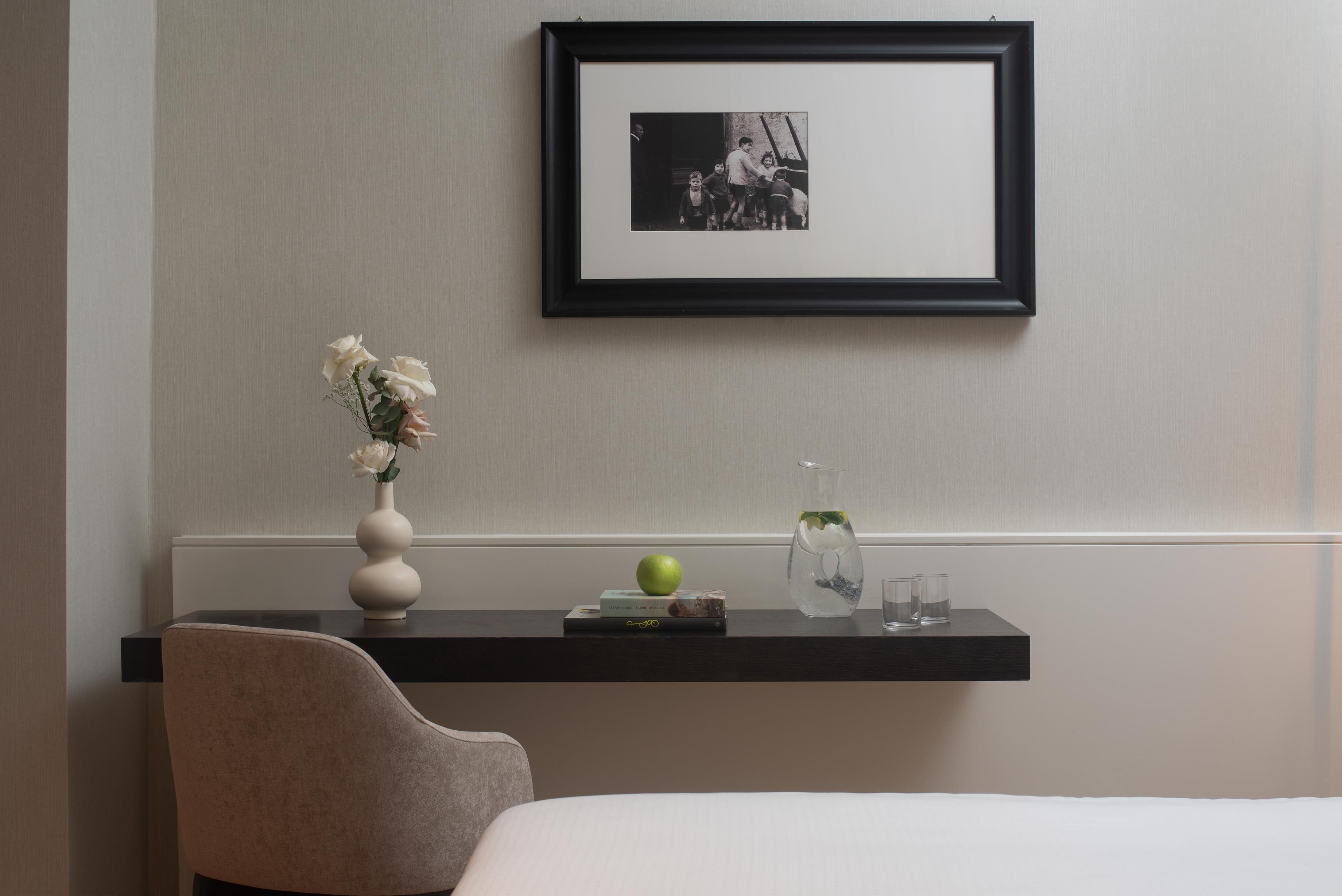 A neatly arranged hotel room with a bed, a chair, a desk with a vase of flowers, a framed photo, a book, and a glass of water.