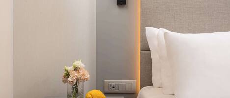 Una habitación de hotel moderna con una mesita de noche, un florero con flores, un limón y una lámpara de pared.