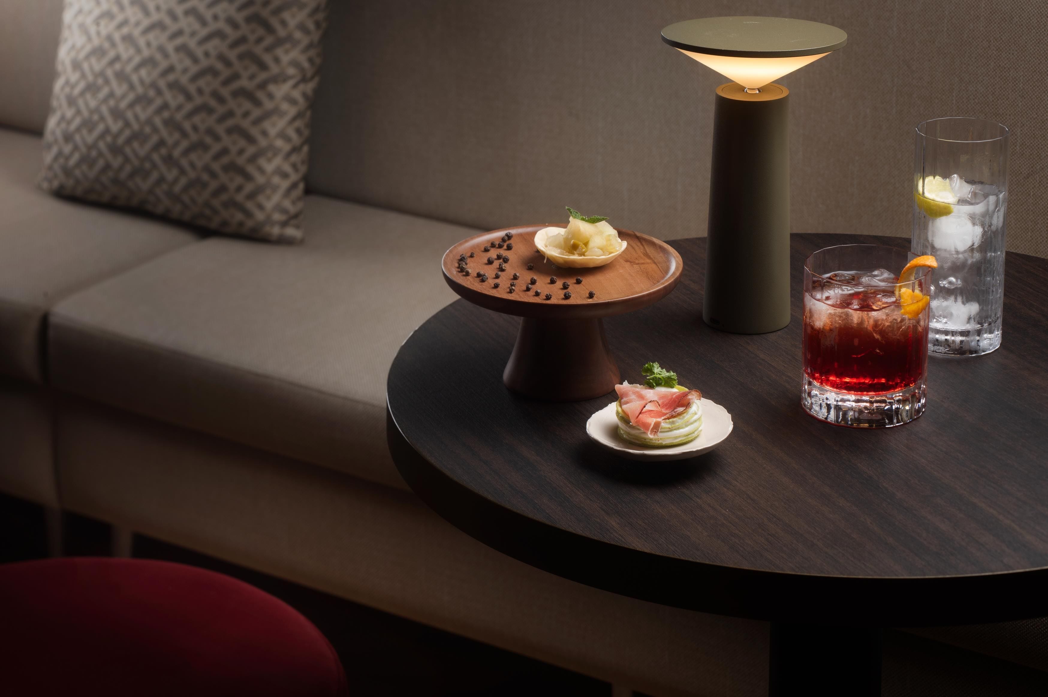 A round table with a lamp, two glasses of iced drinks, and three small plates with appetizers.