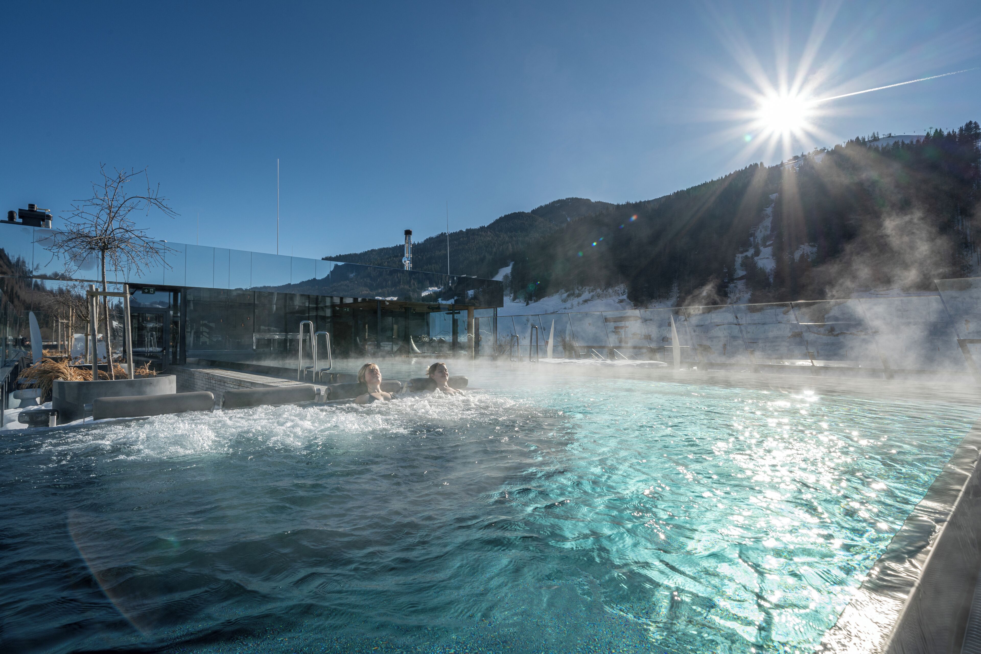 Uma piscina de águas termais com água cristalina, rodeada por um compartimento de vidro e com montanhas ao fundo.
