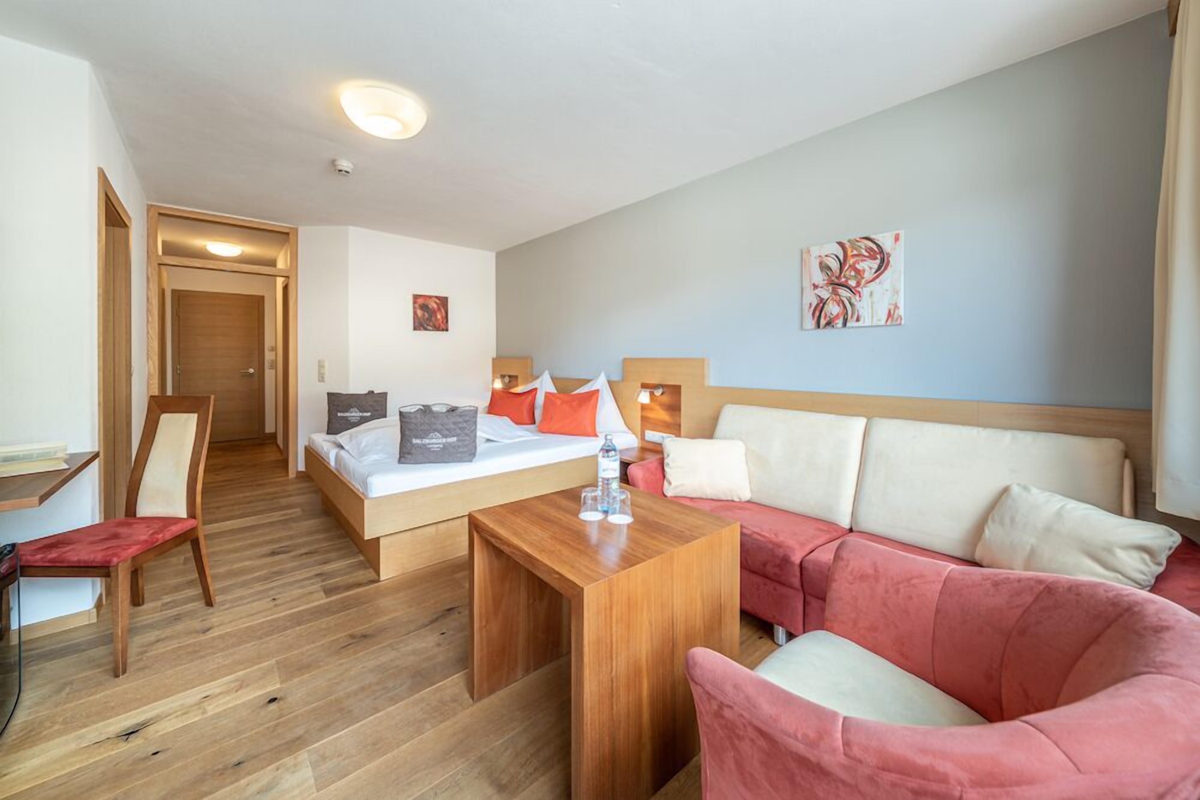 A modern hotel room with a wooden floor, a bed with white linens, a pink sofa, a wooden coffee table, and a desk with a chair.