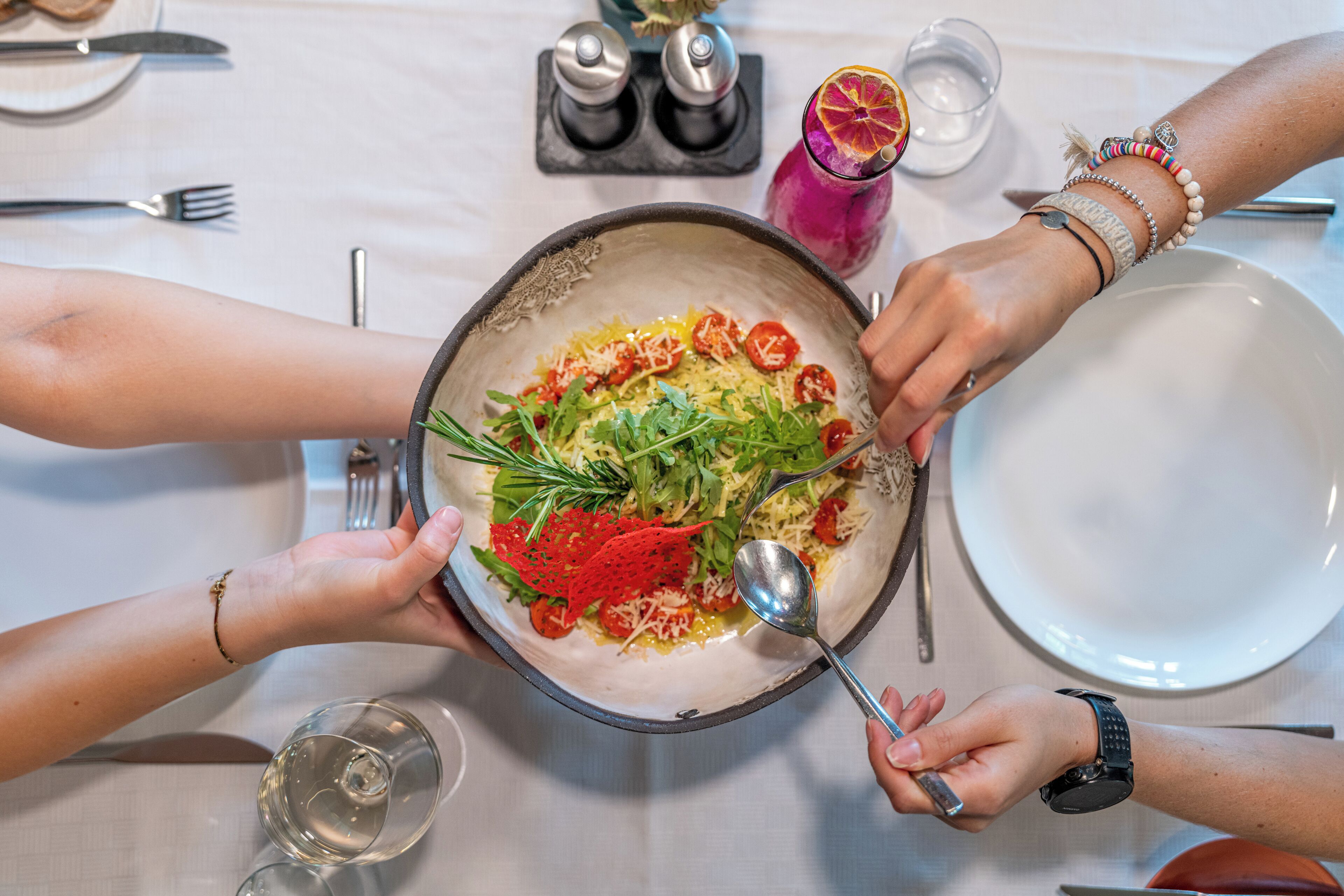 Una mesa de comedor con un bol grande de ensalada, varios utensilios y un vaso de agua.