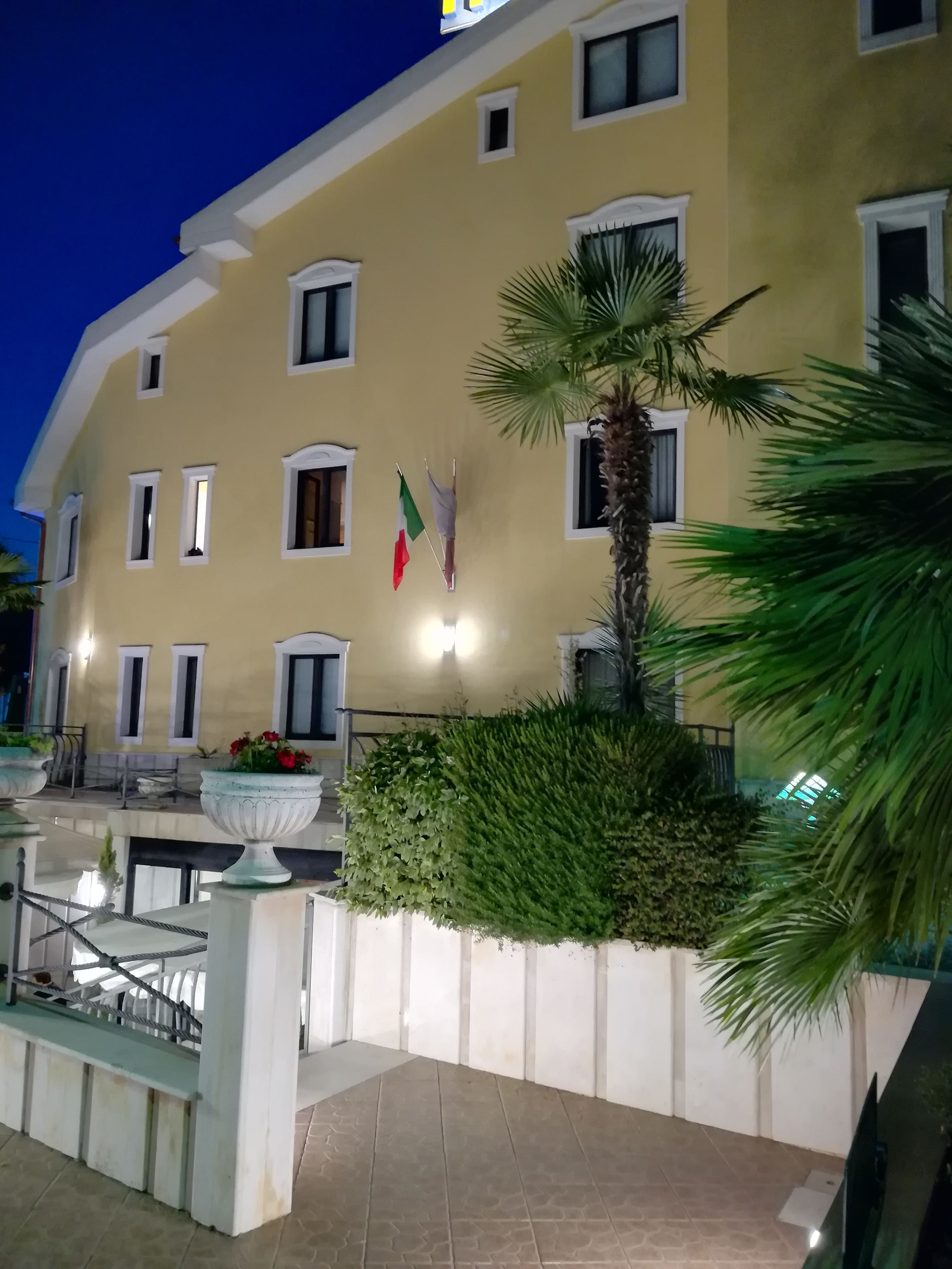 Hotelgevel in de schemering met een gele gevel, meerdere ramen en een verzorgde tuin met een palmboom en bloempotten.