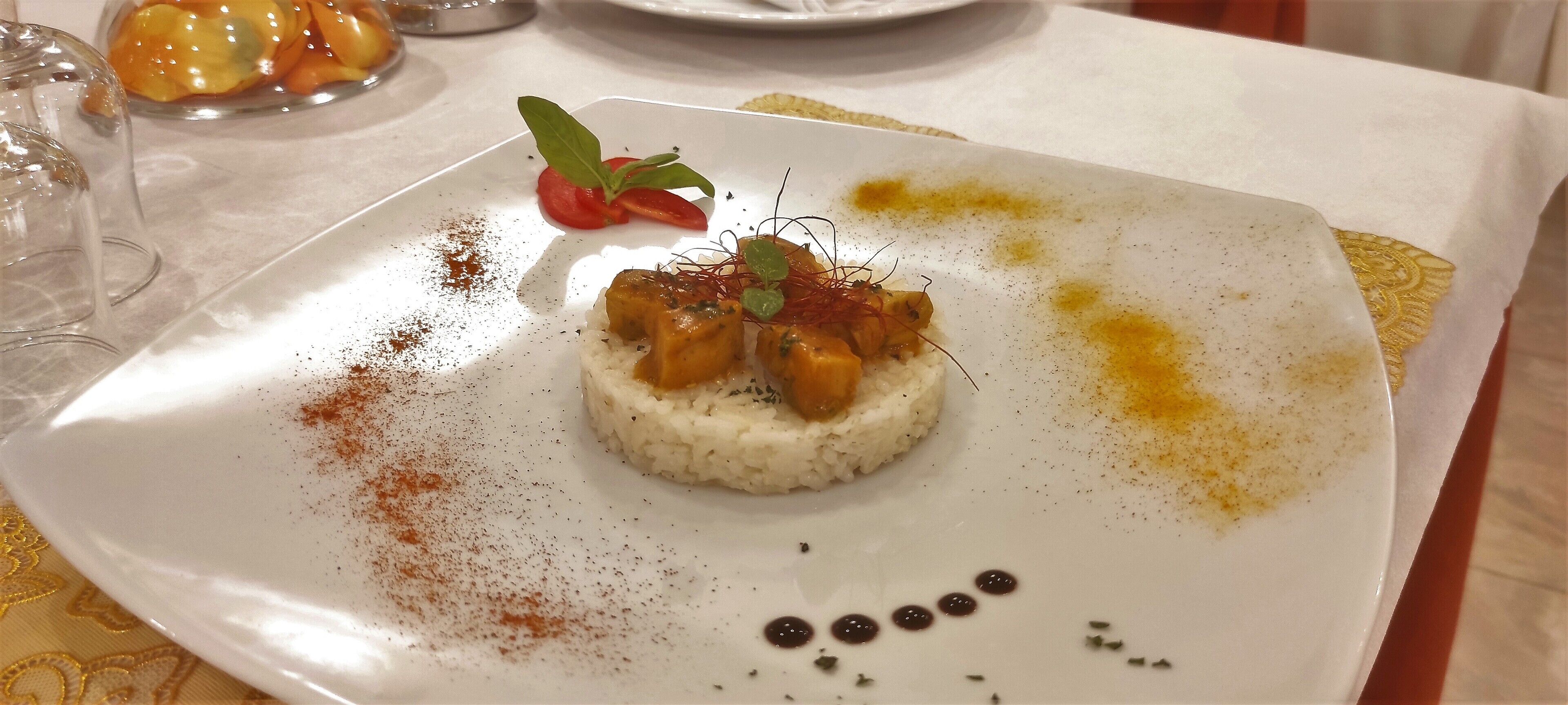 A white plate with a dessert featuring a central mound of rice, topped with a piece of meat and garnished with herbs.