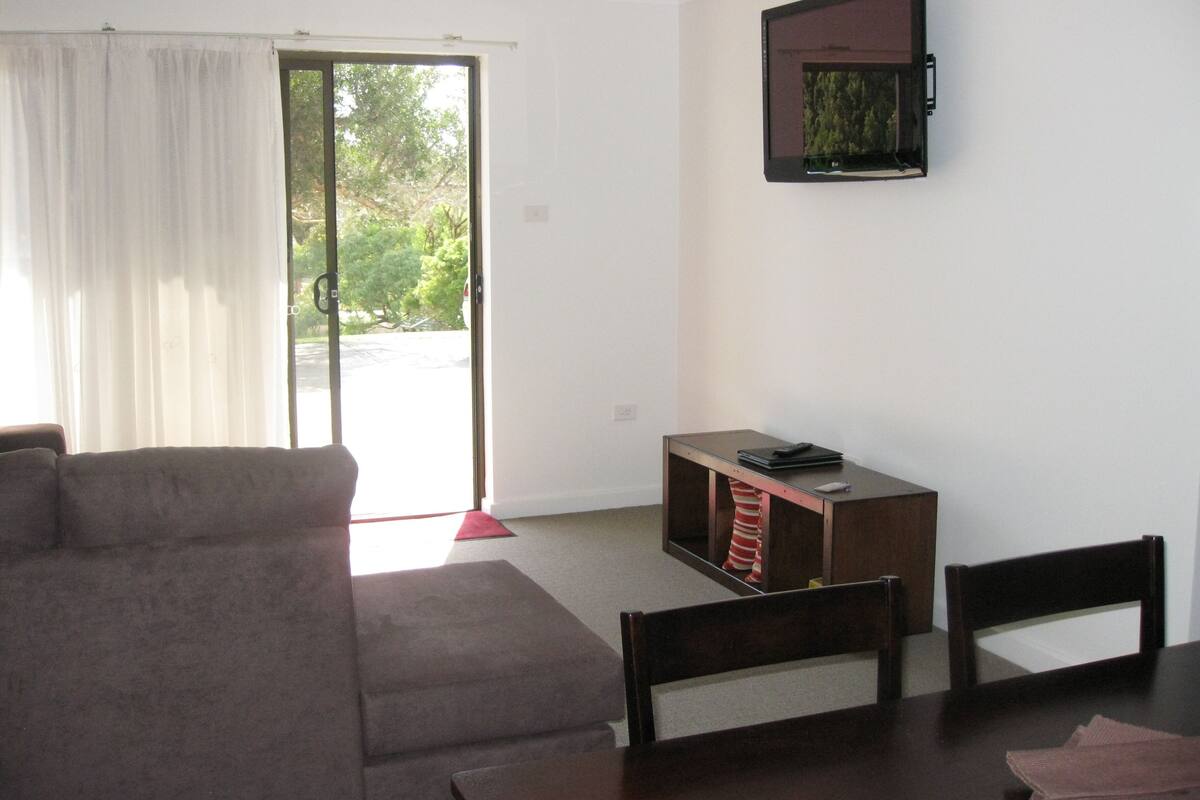 Ein Wohnzimmer mit einer Couch, einem Esstisch, Stühlen, einer kleinen Holzeinrichtung und einem an der Wand montierten Fernseher.