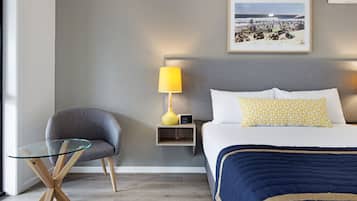 Habitación de hotel con una cama, una mesita de cristal, una silla, una mesita de noche con lámpara y un cuadro en la pared.