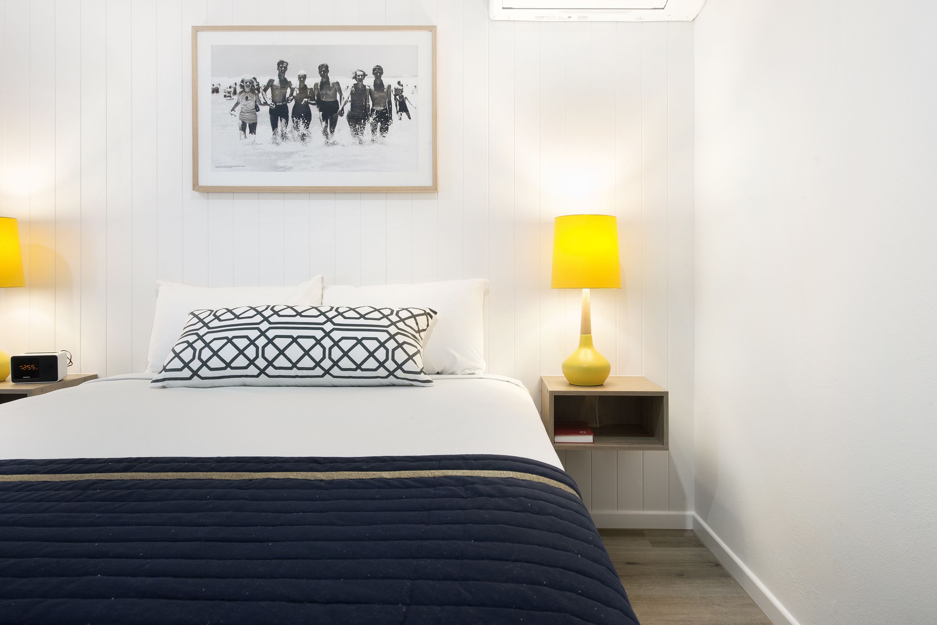 Un letto rifatto con cura, un cuscino decorato, un comodino con una lampada e un quadro appeso al muro.