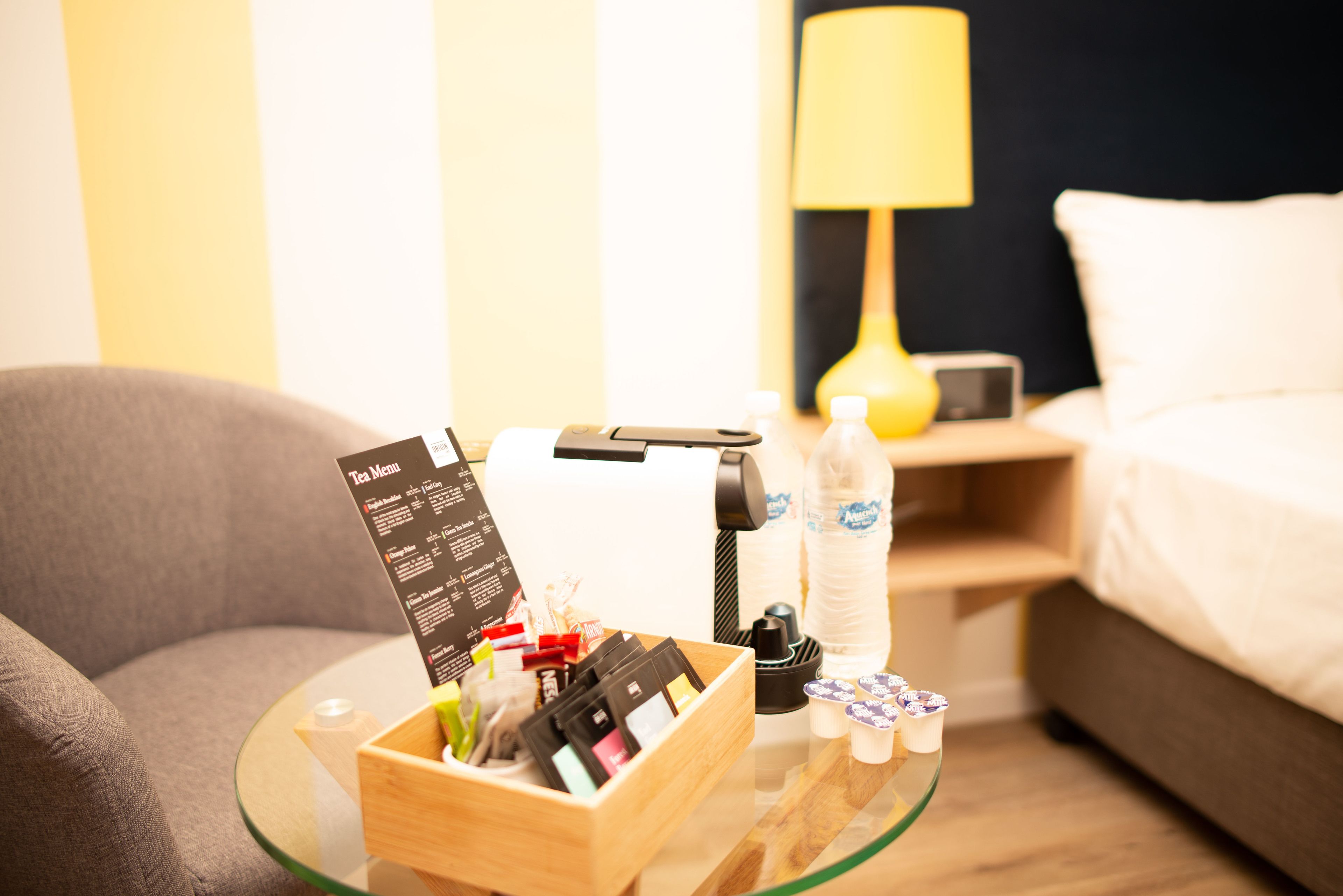 Une chambre d’hôtel avec une table en verre sur laquelle se trouvent un menu de thés, une machine à café et une bouteille d’eau.