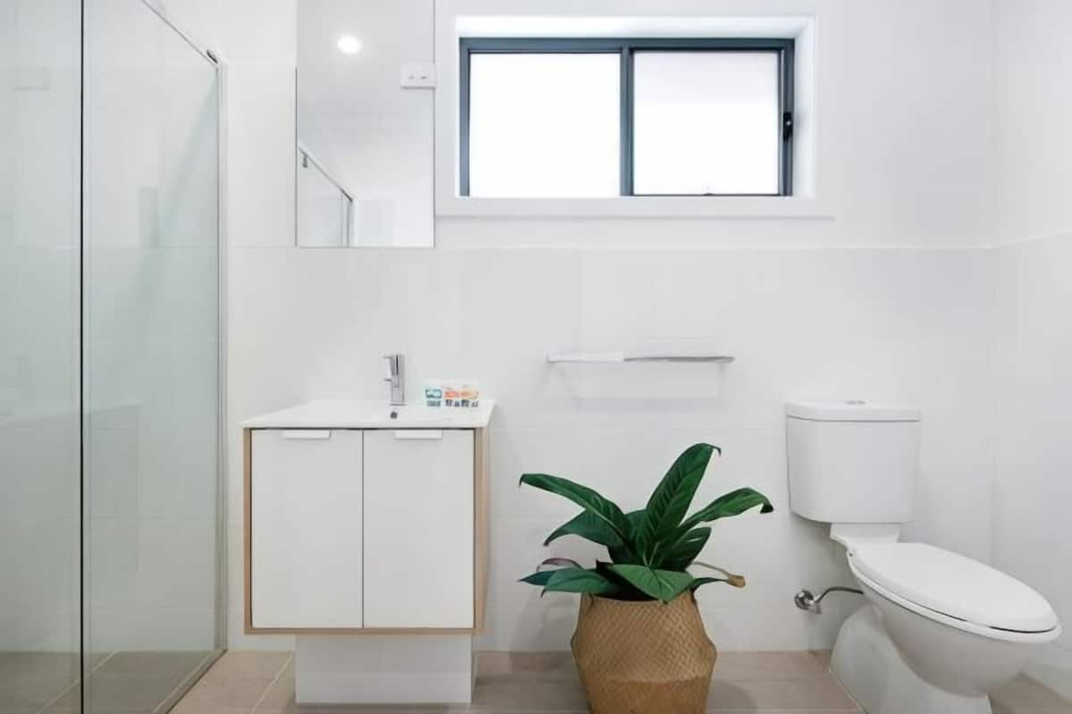 A modern bathroom with a glass shower enclosure, a white sink with a cabinet, a toilet, and a window above the sink.