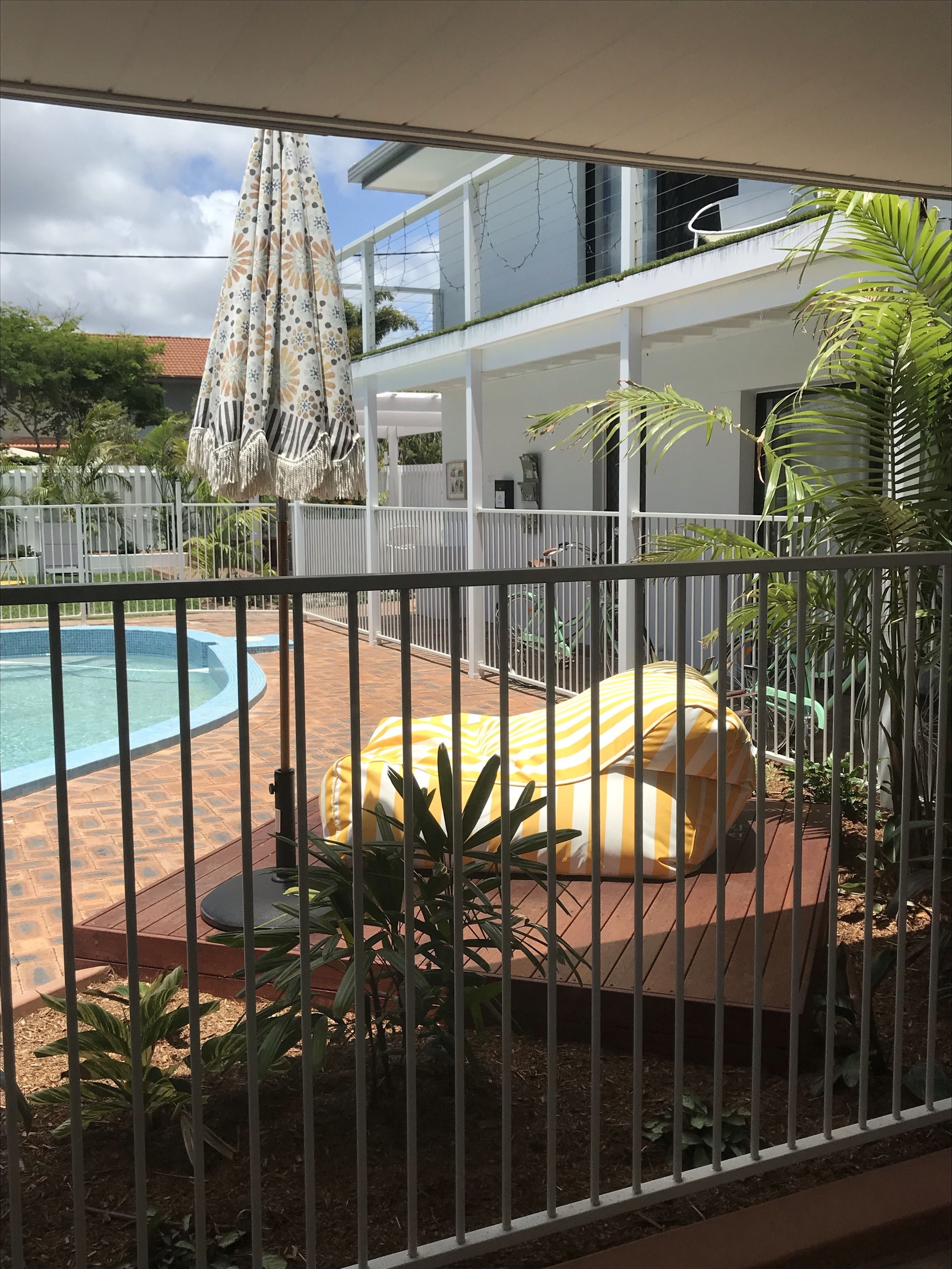 A pool area with a white fence, a yellow-striped lounge chair, and a covered seating area with a patterned umbrella.