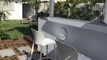 A patio area with two white bar stools, a pergola, and a view of a residential area with palm trees and a house.