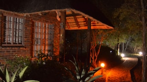 A brick building with a window featuring metal bars, a wooden pergola, and a well-lit garden area with plants and a pathway. - Phumula Kruger Lodge (Nkomazi)