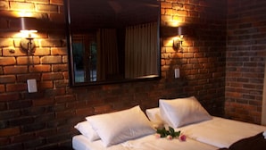 A hotel room with a brick wall, a large bed with white linens, two wall-mounted lamps, and a mirror above the bed. - Phumula Kruger Lodge (Nkomazi)