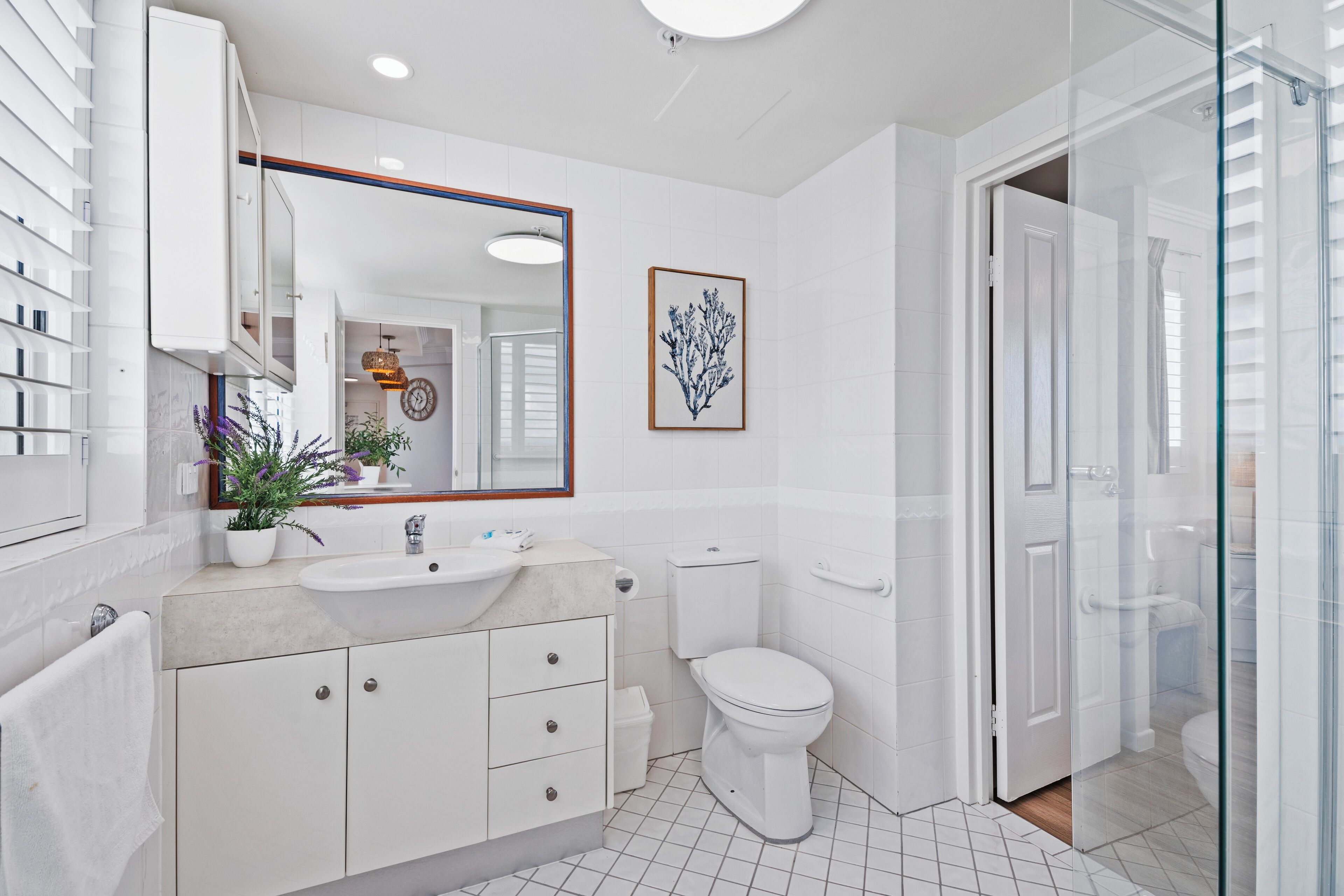 A modern bathroom with a white sink, mirror, and toilet. There is a glass shower enclosure and a potted plant on the counter.
