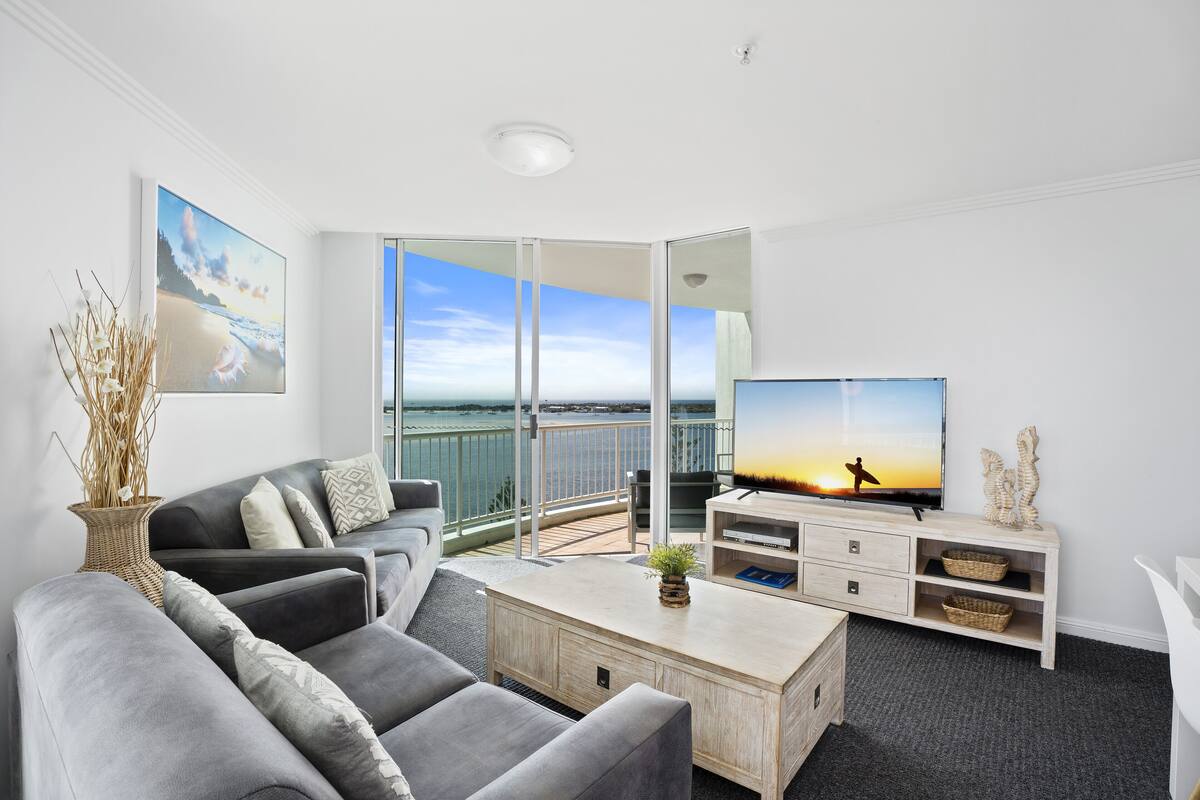 A modern living room with a grey sofa set, a wooden coffee table, and a flat-screen TV. A large window offers a view of the sea and a balcony with a glass railing.