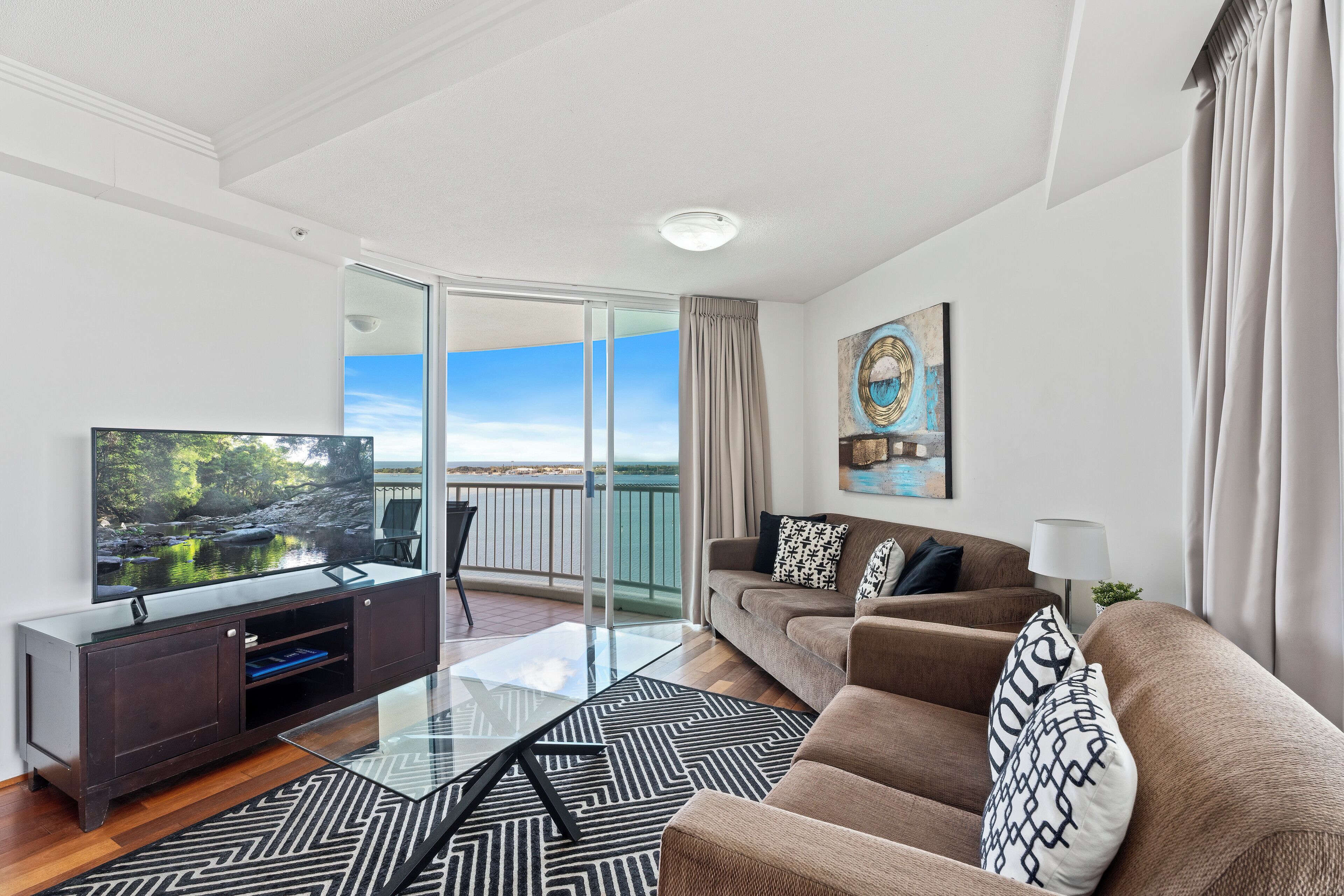A modern living room with a glass coffee table, a sofa, a TV on a cabinet, and a balcony with a view.