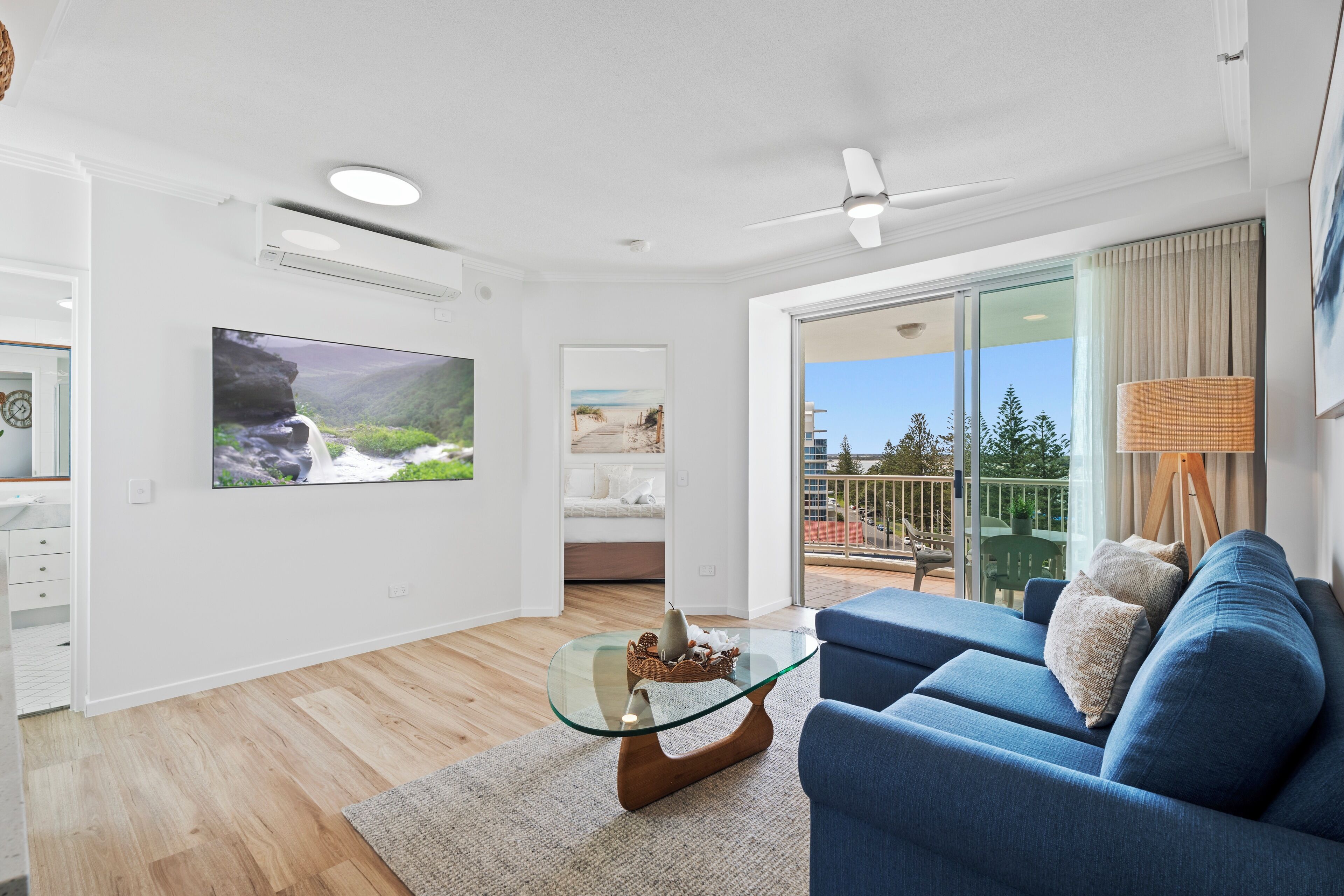 Ett modernt vardagsrum med ett soffbord i glas, en blå soffa, en platt-TV och en balkong med utsikt över staden.