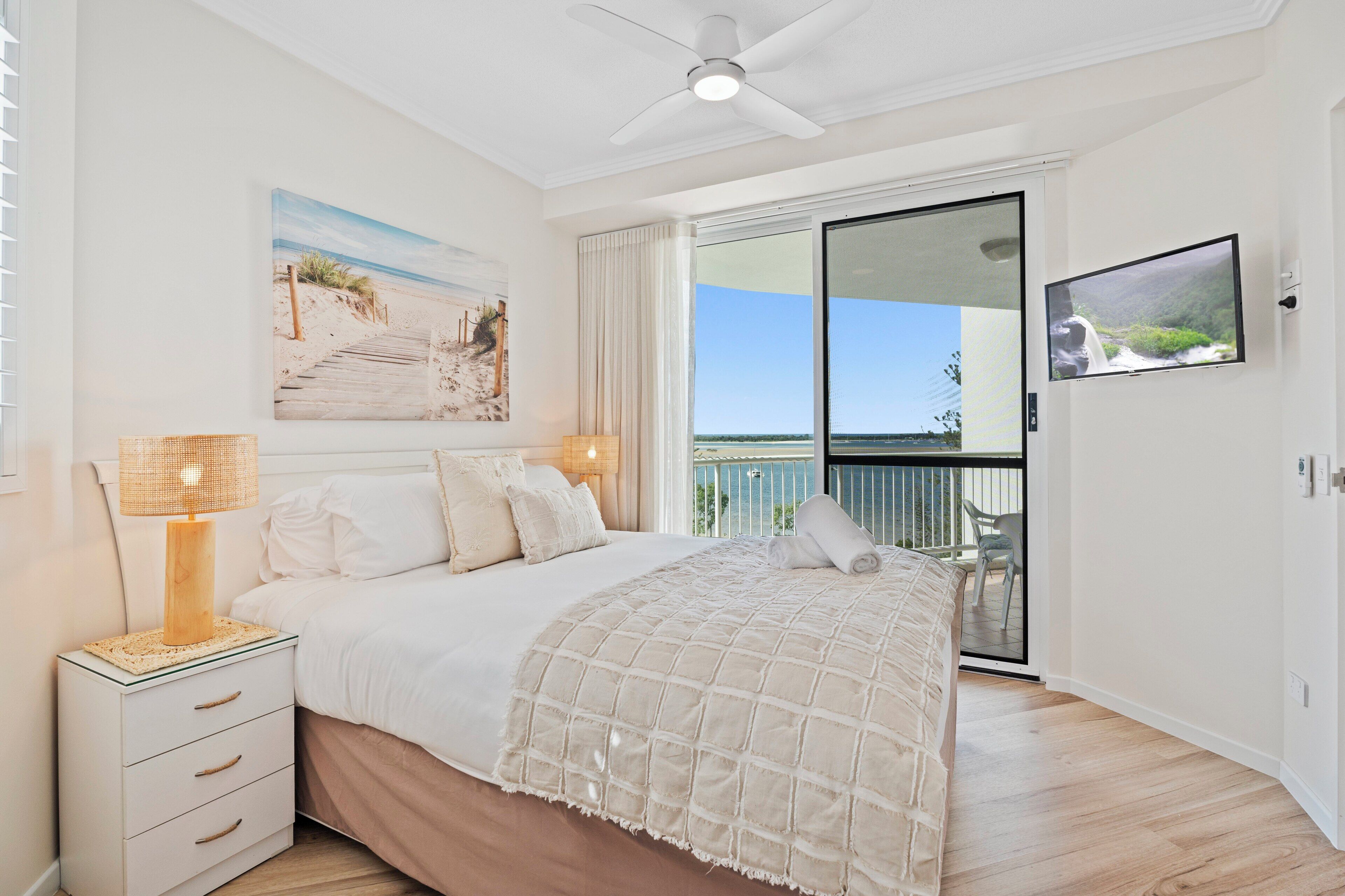 A bedroom with a bed, bedside lamps, a ceiling fan, a large window, and a balcony with a chair.