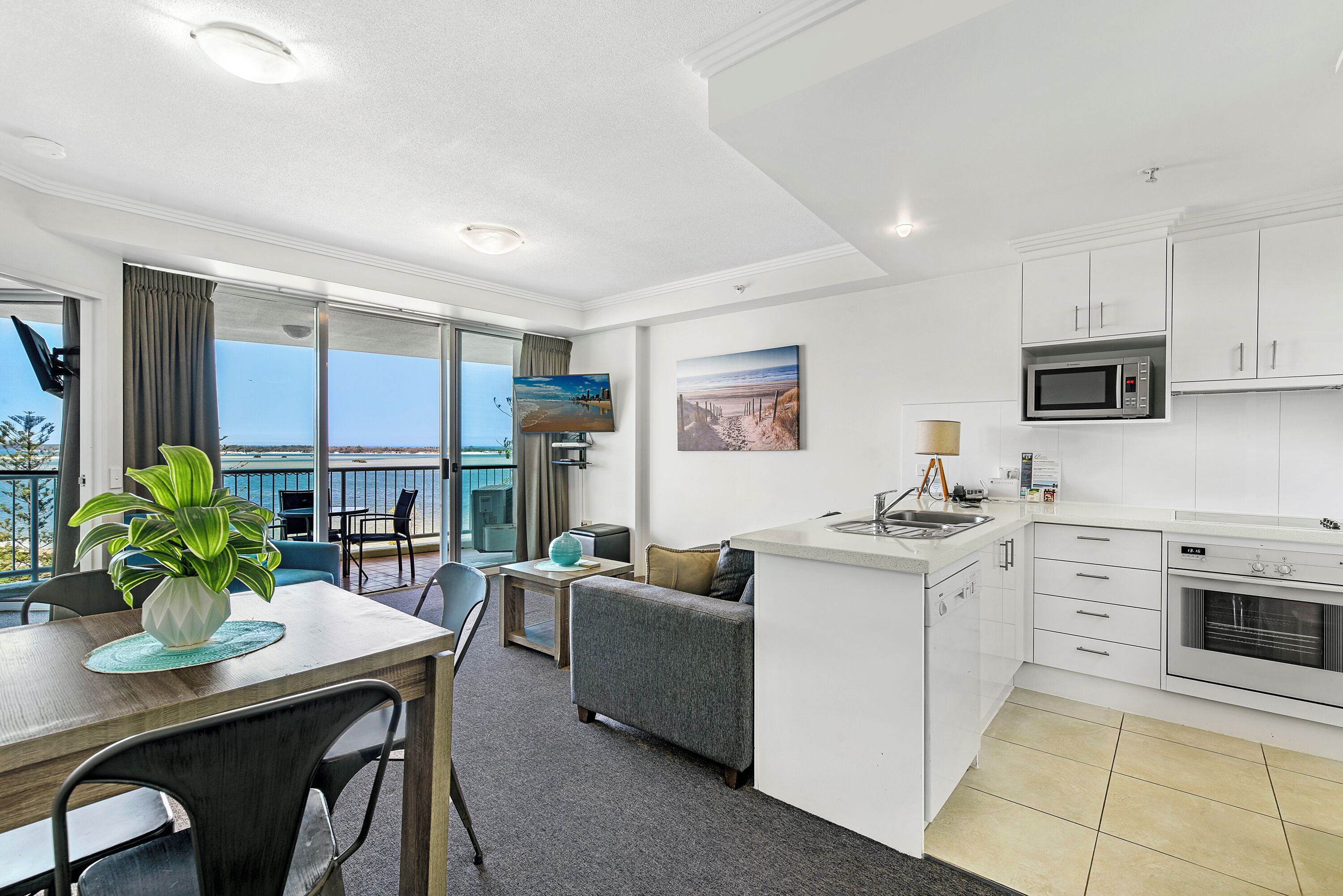 A modern kitchen with white cabinets, a microwave, and a built-in oven. There is a dining area with a table and chairs, and a sofa. A plant is on the table, and there are large windows with a view of the ocean.