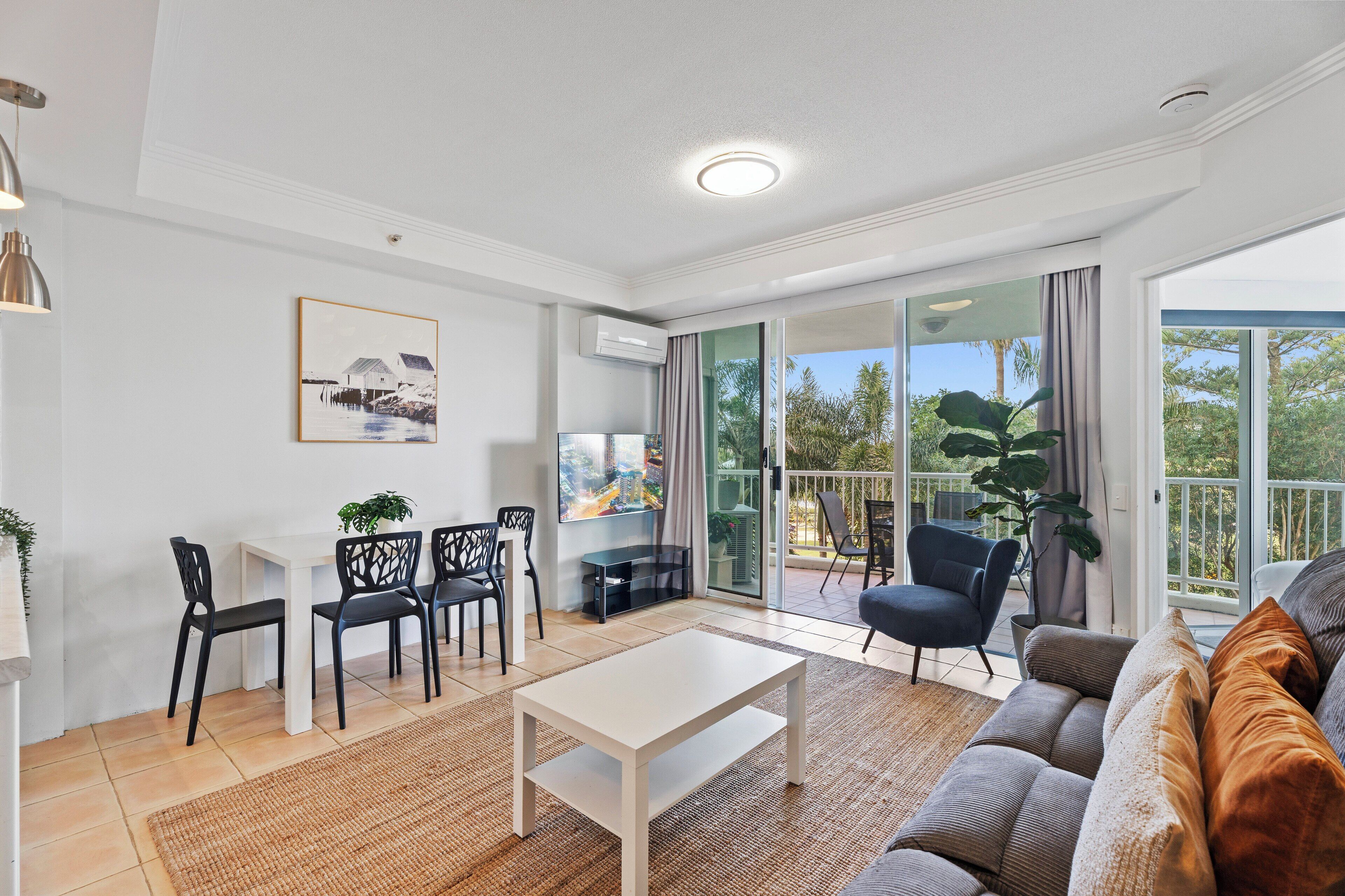 A modern living room with a white coffee table, black chairs, a grey sofa, and a large window with a view of trees.