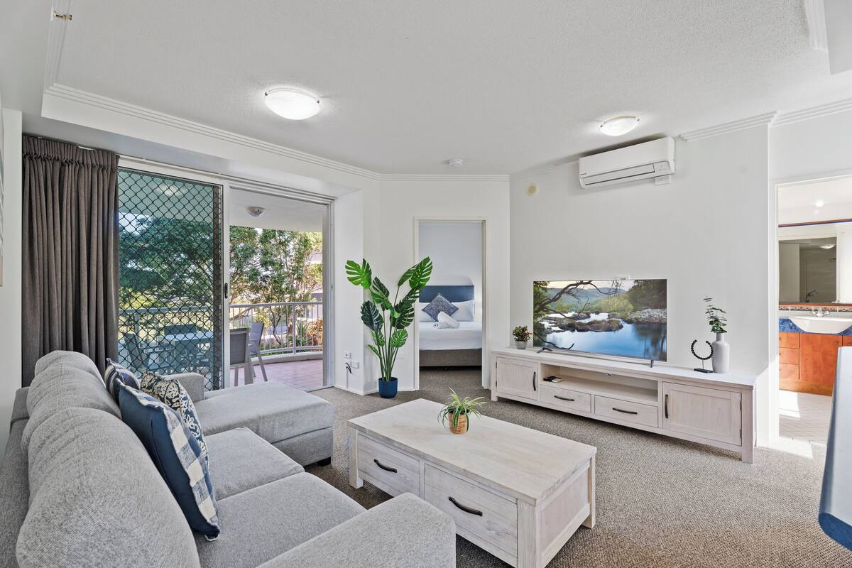 A modern living room with a grey sofa, a wooden coffee table, and a TV. There is a sliding glass door leading to an outdoor area with a view of trees and a pool.