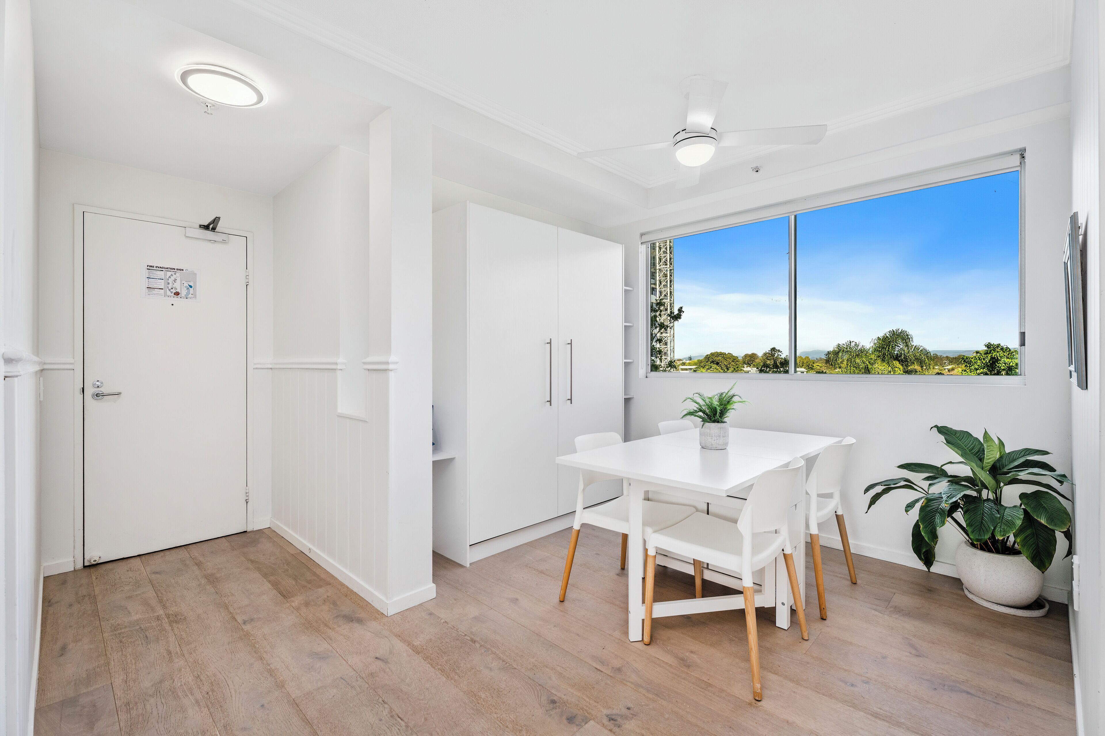 A modern kitchen with white cabinetry, a dining table, and a large window offering a view of greenery and a clear sky.