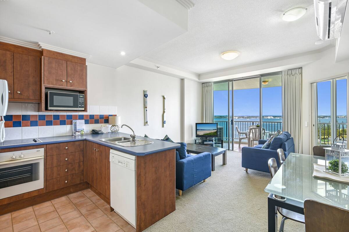 A modern kitchen with a built-in microwave, a sink, and wooden cabinets. There is a dining area with a glass table and chairs, and a balcony with a view of the sea.