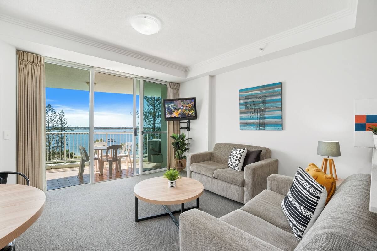 A modern living room with a sofa, armchairs, a coffee table, and a TV. There's a balcony with a view of the sea and trees.