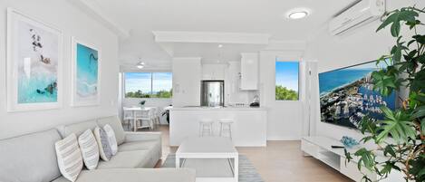 A modern living room with a white sofa, a dining table, and a kitchen area in the background.