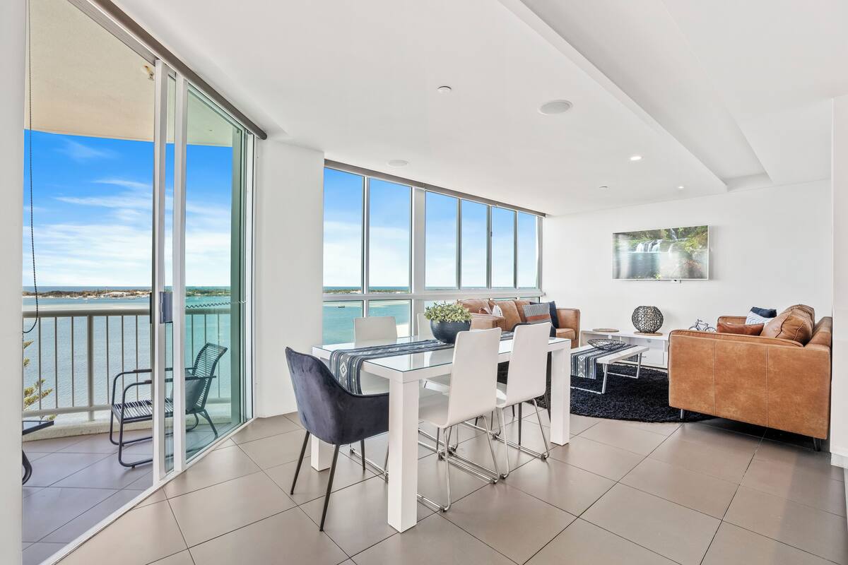 A modern living room with a dining area, large windows, and a balcony overlooking the sea.