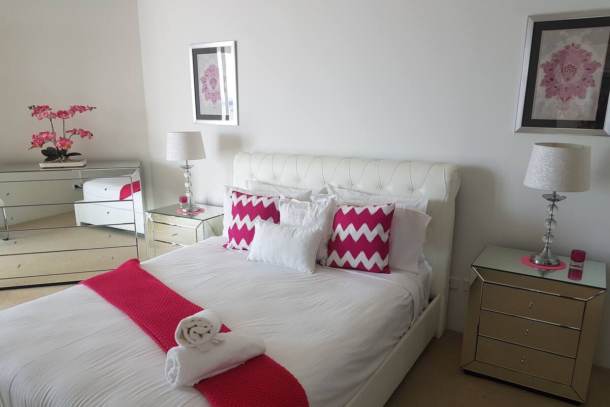 A neatly arranged bedroom with a large bed, white and pink bedding, a bedside table with a lamp, and a dresser.