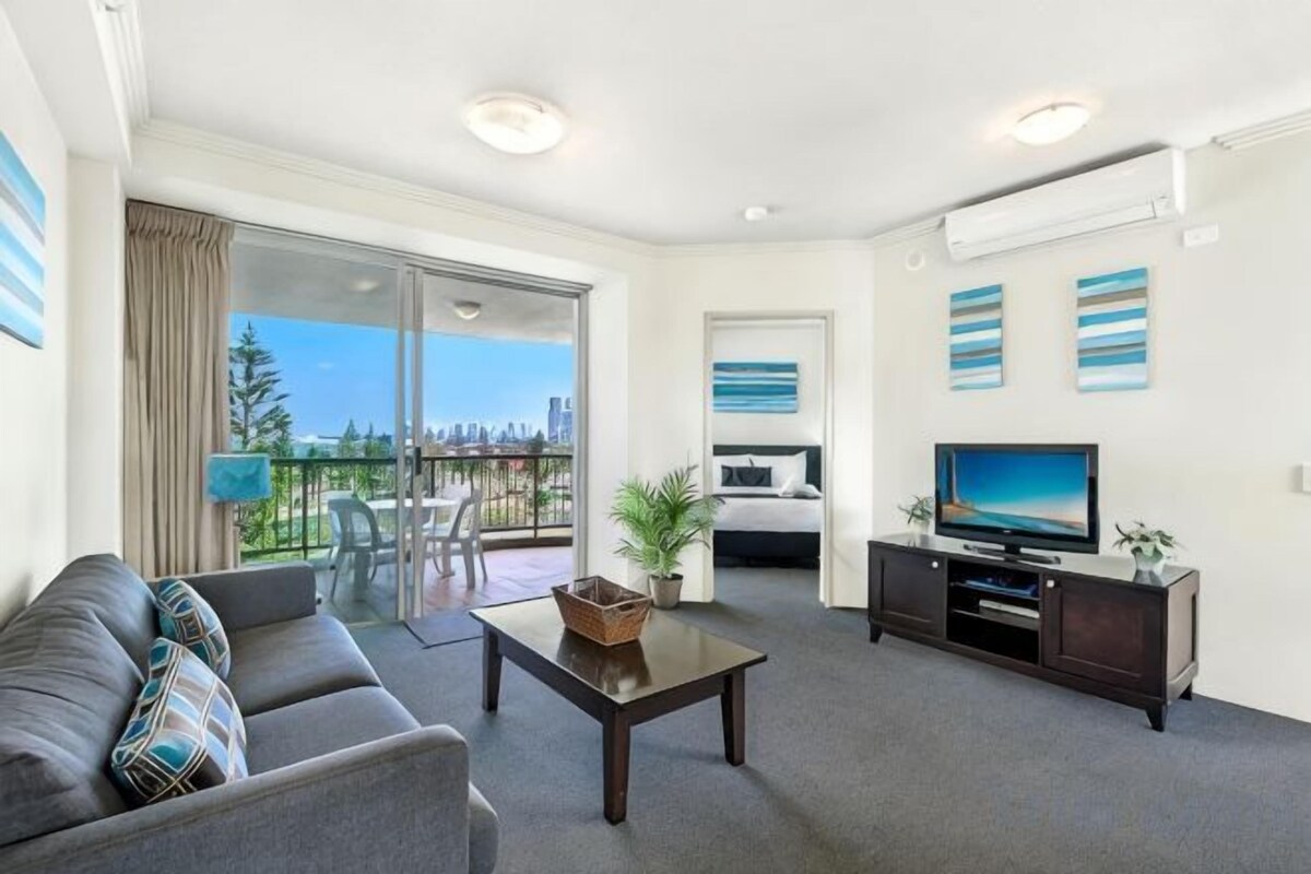 A modern living room with a sofa, a coffee table, a flat-screen TV, and a balcony with a city view.