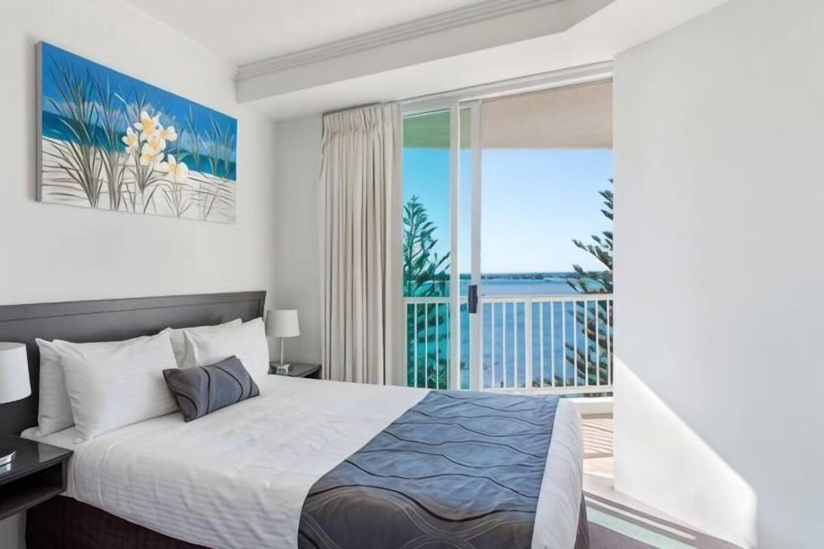 A hotel room with a bed, a nightstand, a painting, and a balcony with a view.