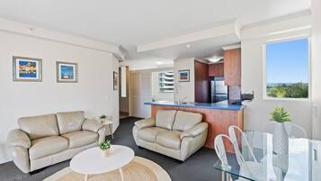 Una sala moderna con dos sofás de cuero color beige, una mesa de centro de vidrio, y un comedor con una mesa y sillas de vidrio. Se ve una cocina con cubierta azul y armarios de madera al fondo.